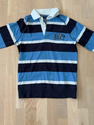 Randig långärmad tröja från Gant  - Säljer en klassisk rugbytröja från Gant med breda blå, marinblå och vita ränder. Skjortan har vit krage, knappar i halsen och GANT-logga broderad på bröstet. Tillverkad i mjuk bomull och har lång ärm.