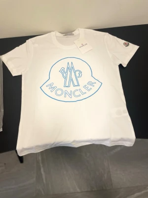Vit Moncler t-shirt med blå logga - Säljer en vit t-shirt från Moncler med stor blå logga framtill och klassisk rund hals. T-shirten har korta ärmar och är tillverkad i mjuk bomull. Moncler-märke på ärmen för extra detalj. Perfekt för dig som gillar stilrena och exklusiva plagg.