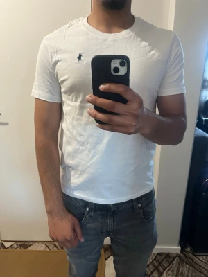 Vit t-shirt från Polo Ralph Lauren - Klassisk vit t-shirt från Polo Ralph Lauren med den ikoniska loggan broderad på bröstet. T-shirten har rund hals, korta ärmar och är gjord i mjuk bomull för en skön känsla. Perfekt basplagg som funkar till allt.