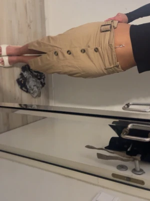 Beige kjol med knappar och bälte - Säljer en beige kjol i bomull med knappar framtill och ett matchande bälte i midjan. Kjolen har en slits framtill som ger en snygg detalj och är perfekt för vår och sommar. Enkel att styla med både sneakers och sandaler.