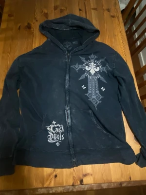 Svart hoodie från Affliction - Svart hoodie med dragkedja från Affliction. Tryck med kors, dödskallar och texten 'Live Fast' både fram och bak. Stora fickor framtill och huva. Snygg streetwear-stil med slitna detaljer och unika grafiska prints.