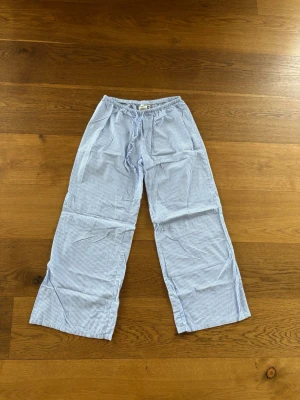 Randiga pyjamasbyxor från H&M - Säljer ett par klassiska pyjamasbyxor från H&M i ljusblått och vitt med tunna ränder. Byxorna har resår i midjan och är tillverkade i mjuk bomull, perfekta för chill dagar hemma eller mysiga kvällar.