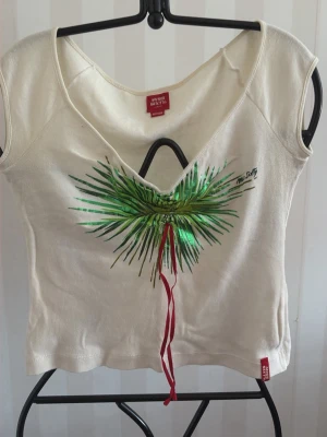 Vit topp med palmtryck från Miss Sixty - Vit kortärmad topp från Miss Sixty med grönt palmbladstryck och röd snörning framtill. V-ringad hals och figurnära passform. Tillverkad i mjuk bomull, perfekt för en avslappnad och trendig look.