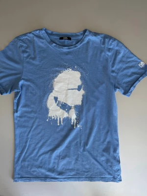 Karl Lagerfeldt T-hir - Blå t-shirt från Karl Lagerfeld med ett stort vitt tryck av Karl Lagerfelds ikoniska siluett på bröstet. T-shirten har korta ärmar och rund hals. Nypris : ca 700 SEK Pris : 249 SEK