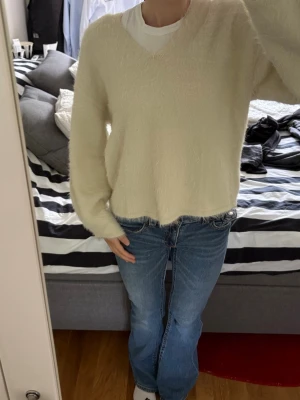 Krämvit fluffig stickad tröja - Mysig krämvit stickad tröja med v-ringning och lång ärm. Tröjan har en fluffig textur och avslappnad passform, perfekt att bära över en t-shirt. Passar till jeans för en chill look.
