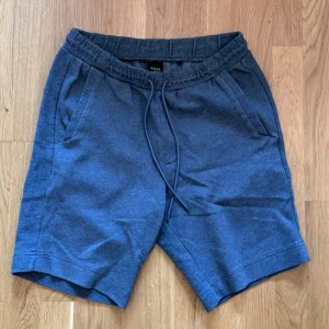 Hugo Boss Shorts - Snygga grå shorts från Hugo Boss med elastisk midja och snörning framtill. Shortsen har två sidofickor och är tillverkade i mjuk bomull, perfekta för en avslappnad stil. Boss-logga diskret broderad på sidan.