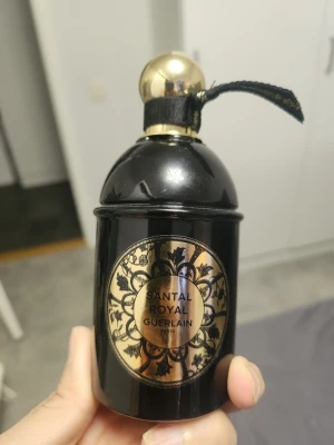 Santal Royal parfym från Guerlain - Lyxig parfymflaska från Guerlain, Santal Royal. Flaskan är svart med gulddetaljer och har en rundad form med elegant guldlock och svart band. Märkets logga och namn syns på en dekorativ etikett framtill. Perfekt för dig som gillar exklusiva dofter och snygg design.