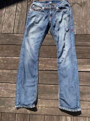 Blå jeans med röd kontrastsöm - Säljer ett par blå jeans med raka ben och snygga slitningar. Jeansen har vita och röda kontrastsömmar samt detaljerade fickor med silverfärgade knappar. Materialet är klassisk denim i bomull. Perfekta för en avslappnad och trendig look.