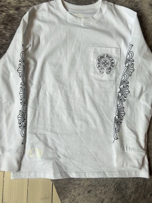 Vit långärmad topp från Chrome Hearts - Säljer en vit långärmad topp från Chrome Hearts med svart grafiskt tryck längs ärmarna och logga på bröstfickan. Toppen har rund hals och är tillverkad i mjuk bomull. Perfekt för dig som gillar streetwear och snygga detaljer.