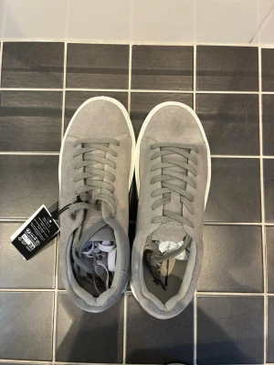 Gråa sneakers från Jack & Jones - Stilrena gråa sneakers från Jack & Jones i mockaimitation med matchande grå skosnören och vit sula. Klassisk låg modell med enkel design som passar till det mesta. Perfekta för dig som gillar en clean och modern look.
