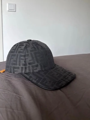 Svart Fendi keps med logomönster - Säljer en svart keps från Fendi med det klassiska FF-logomönstret över hela kepsen. Kepsen har böjd skärm och en liten gul detalj på sidan. Tillverkad i tygmaterial som känns både stilrent och lyxigt.