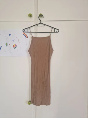 Beige ribbad midiklänning med smala band - Snygg beige ribbad klänning med smala axelband och rak passform. Klänningen har en enkel design med rundad rygg och är tillverkad i ett stretchigt material som sitter skönt på kroppen. Perfekt för varma dagar och enkel att styla.