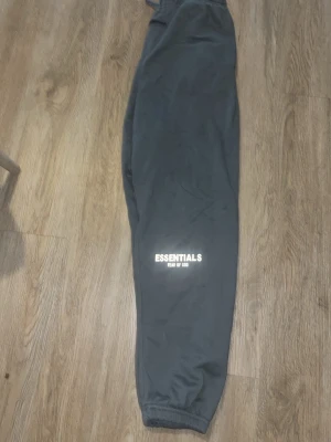 Gråa Essentials mjukisbyxor - Mörkgrå mjukisbyxor från Fear of God Essentials med tryckt logga på benet. Byxorna har resår i midjan och vid bensluten samt en bakficka. Tillverkade i mjukt material för maximal komfort och avslappnad passform.