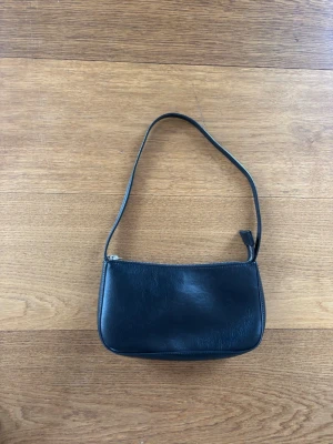Svart axelremsväska i skinn Brandy Melville - En stilren svart handväska i slätt skinn med enkel axelrem och dragkedja upptill. Väskan har en minimalistisk design och en klassisk, rektangulär form som passar till många olika looks.