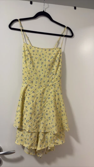 Gul blommig jumpsuits - Söt gul kort jumpsuits med tunna axelband och blått blommönster. Klänningen har volangdetaljer i kjolen och en lätt, luftig känsla som passar perfekt till varma dagar. Materialet känns mjukt och bekvämt mot huden.