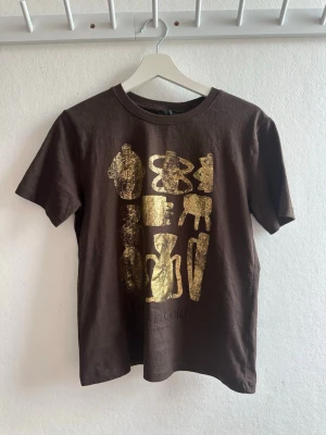 Brun t-shirt med guldtryck - Snygg brun t-shirt med korta ärmar och ett unikt guldmönster framtill. T-shirten har en klassisk passform och är tillverkad i mjuk bomull, perfekt för en avslappnad och trendig stil.