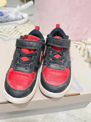 Nike svart-röda sneakers med kardborre - Snygga sneakers från Nike i svart och rött med klassisk design. Skorna har kardborreband, svarta skosnören och vit sula. Ovandelen är i läder och mesh för bra andningsförmåga. Perfekta för dig som gillar sportig stil och vill sticka ut.