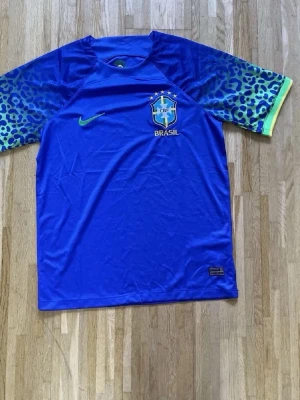 Blå Brasilien Nike träningströja - Snygg blå träningströja från Nike med Brasilien-logga på bröstet. Ärmarna har ett leopardmönster i blått och grönt med gul kant. Tillverkad i lätt och snabbtorkande material, perfekt för fotbollsträning eller gymmet.