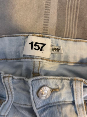 Ljusblå flare jeans från 157 - Säljer ett par ljusblå flare jeans från 157 med hög midja och klassisk femficksdesign. Jeansen har en snygg knappdetalj framtill och är tillverkade i ett mjukt denimtyg som ger en skön passform. Perfekta för dig som gillar retrovibbar och vill sticka ut.