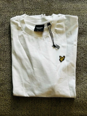 Vit t-shirt från Lyle & Scott - Klassisk vit t-shirt från Lyle & Scott med rund hals och korta ärmar. T-shirten har den ikoniska gula logon broderad på bröstet och är tillverkad i mjuk bomull för en skön känsla. Perfekt basplagg med stilren design.