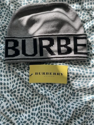 Grå mössa från Burberry - Snygg grå mössa från Burberry med stor svart logotyp framtill. Tillverkad i mjukt stickat material som känns skönt mot huden. Klassisk beanie-modell som passar perfekt till streetwear och ger en exklusiv touch till din outfit.