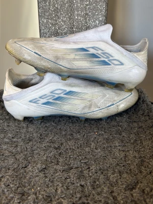 Vita Adidas F50 fotbollsskor - Säljer ett par vita Adidas F50 fotbollsskor med blå detaljer och stilren design. Skorna har låg profil, smala dobbar och är tillverkade i syntetmaterial för lätt vikt och bra bollkontroll. Perfekta för snabba spelare som vill sticka ut på planen. Skorna är i jättebra skick men är bara smutsiga. Pris kan diskuteras