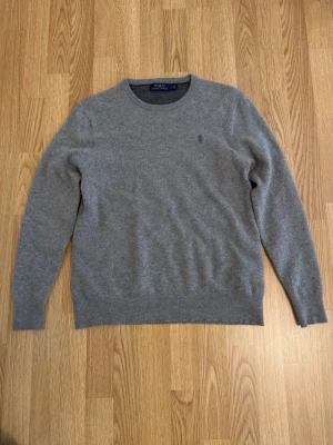 Grå stickad tröja från Polo Ralph Lauren - Klassisk grå stickad tröja från Polo Ralph Lauren med rund halsringning och diskret broderad logga på bröstet. Hör av dig vid fler frågor.