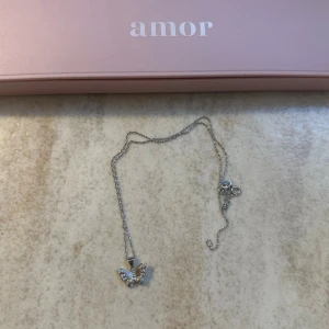 Silverhalsband med fjäril från Amor - Snyggt silverfärgat halsband från Amor med en detaljrik fjärilsberlock. Kedjan är tunn och smidig, och fjärilen har små glittrande stenar som ger extra bling. Perfekt accessoar för dig som gillar söta och trendiga smycken.