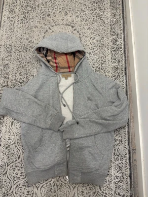 Grå hoodie från Burberry med dragkedja - Snygg grå hoodie från Burberry med klassiskt rutigt foder i huvan och dragkedja framtill. Varför jag säljer så billigt eftersom dragkedjan funkar inte men den går att fixa