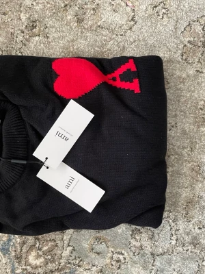  Ami Paris tröja - Svart stickad tröja från Ami Paris med ikoniskt rött hjärta och A på bröstet. Tröjan har rund halsringning, ribbade muddar och är tillverkad i mjukt material. Perfekt för dig som gillar stilrena plagg med en twist.
