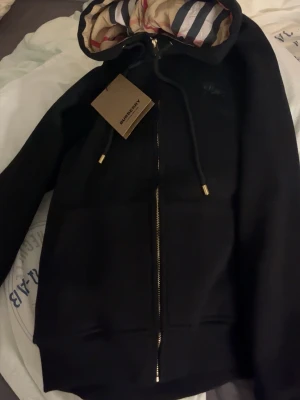 Svart Burberry zipup - Säljer en svart hoodie från Burberry med klassisk dragkedja och huva. Insidan av huvan har Burberrys rutmönster i beige, rött och blått. Guldiga detaljer på snören och dragkedja. Lappen är inte kvar men den är knappast använd så den är som ny.