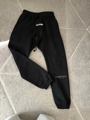  Essentials mjukisbyxor - Svarta mjukisbyxor från Essentials Fear of God med logga på vänster ben. Byxorna har resår i midjan, dragsko och muddar vid benslut. Materialet är mjukt och bekvämt, perfekt för chill eller streetwear. Klassisk passform och stilren design.