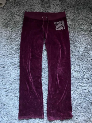 Vinröda velourbyxor Juicy Couture - Mysiga vinröda mjukisbyxor från Juicy Couture i velour. Byxorna har resårmidja med dragsko och texten 'CHOOSE JUICY' i strass på ena höften. Byxorna har en liten defekt på baksidan av midjan (bild finns) 