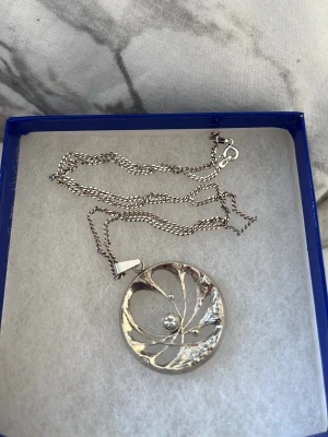 Silverfärgat halsband med rund hängsmycke - Säljer ett silverfärgat halsband med ett runt hängsmycke som har ett snirkligt mönster och en liten pärla i mitten. Kedjan är tunn och har ett klassiskt lås. Perfekt för dig som gillar stilrena och unika accessoarer. Kedjan är i rikigt silver då de står 925 ingraverat. Längden på halsbandet är 47cm långt.