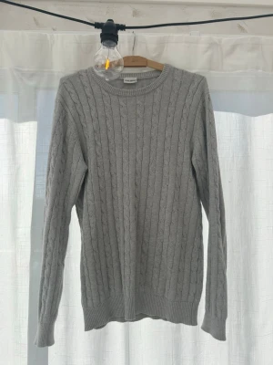 Grå kabelstickad tröja John Henric - Snygg grå kabelstickad tröja från John Henric. Tröjan har rund halsringning, långa ärmar och ribbade muddar vid ärmslut och nederkant. Perfekt för lager-på-lager och enkel att matcha med jeans eller chinos. Ops, lite smutsig på ett ställe 