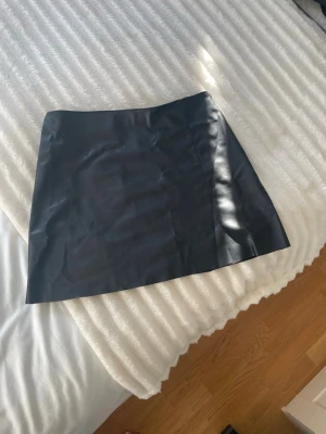 Svart skinnimitation minikjol - Snygg svart kort kjol i skinnimitation. Kjolen har en rak passform och har en dragkedja på sidan. Passar till många olika outfits och är ett måste i garderoben. 