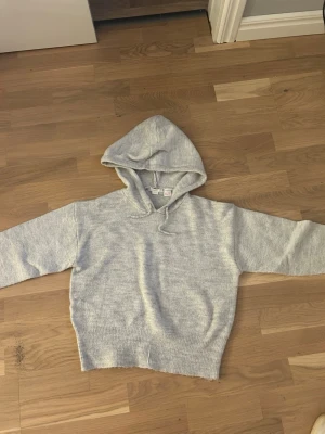 Ljusgrå stickad hoodie - Mysig ljusgrå stickad hoodie med huva och dragsko. Tröjan har en avslappnad passform, långa ärmar och bred ribbad mudd vid midjan. Perfekt för en chill och avslappnad stil.