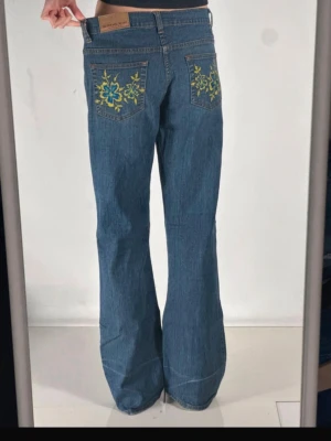 Blå bootcut jeans med broderi - Snygga blå bootcut jeans med broderade blommor i grönt och blått på bakfickorna. Klassisk femficksmodell i denim med låg midja och utsvängda ben. Perfekta för dig som gillar en retro och unik stil. Köpta på Vinted men passade tyvärr inte, bilder från tidigare säljaren.