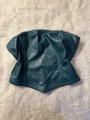 Mörkgrön skinn korsett-topp - Snygg mörkgrön korsett-topp i skinnimitation med en cool, strukturerad form. Toppen är axelbandslös och har en stilren design som ger en edgy vibe. Perfekt för att lyfta vilken outfit som helst.