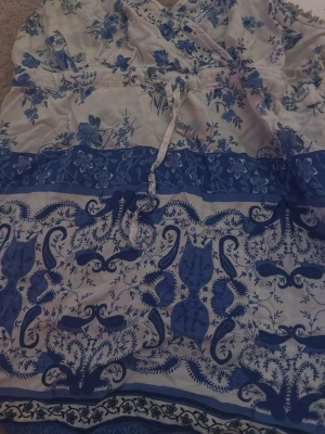 Blåvit blus med blommönster  - Säljer en blåvit blus storlek M. Blusen har ett vackert blommönster och dekorativa band framtill. Materialet är lätt och luftigt, perfekt för varma dagar. Den har en avslappnad passform och fina detaljer i blått och vitt.