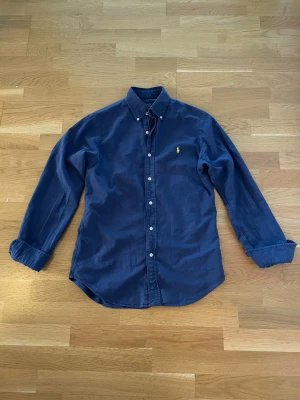 Blå skjorta från Polo Ralph Lauren - Klassisk blå skjorta från Polo Ralph Lauren i slim fit-modell. Skjortan har långa ärmar, knappar framtill och den ikoniska broderade loggan på bröstet. Perfekt för en stilren och avslappnad look.