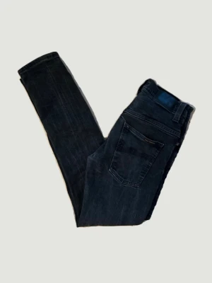 Svarta jeans från Nudie Jeans - Säljer ett par svarta jeans från Nudie Jeans med klassisk femficksdesign och raka ben. Jeansen har en snygg tvättad look och är tillverkade i bomull med lite stretch för extra komfort. Perfekt för dig som gillar en stilren och modern look.