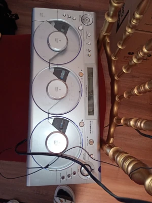 Silver CD-spelare med tre skivfack - Säljer en silverfärgad CD-spelare från märket Sanyo med tre separata CD-fack och digital display. Designen är avlång och stilren med tydliga knappar för kontroll och genomskinliga lock över varje CD-fack. Perfekt för dig som gillar att lyssna på musik från CD-skivor.