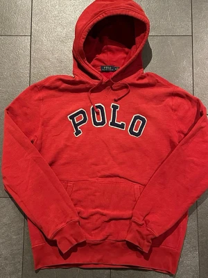 Röd hoodie från Polo Ralph Lauren - Röd hoodie från Polo Ralph Lauren med stor POLO-text i blått och vitt på bröstet. Klassisk känguruficka framtill, justerbar huva med dragsko och ribbade muddar. Perfekt för en avslappnad och sportig stil.