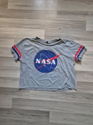 Grå NASA t-shirt med tryck - Cool grå t-shirt med stort NASA-tryck på bröstet och röda samt blå ränder på ärmarna. T-shirten har en avslappnad passform och är gjord i mjuk bomull. Perfekt för dig som gillar rymden och vill ha en unik look.