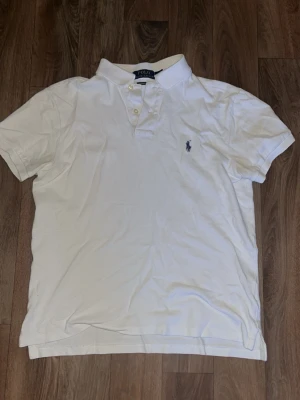 Vit pikétröja från Ralph Lauren - Klassisk vit pikétröja från Ralph Lauren med korta ärmar och krage. Tröjan har knappar framtill och den ikoniska broderade loggan på bröstet. Perfekt för en stilren och avslappnad look.
