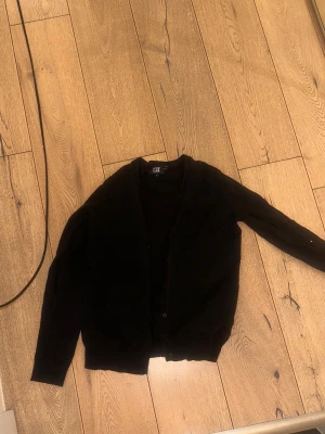 Svart kofta - En klassisk svart kofta med knäppning framtill. Tröjan har lång ärm och ribbade muddar vid ärmslut och nederkant. Perfekt att slänga över en t-shirt eller skjorta för en stilren look.