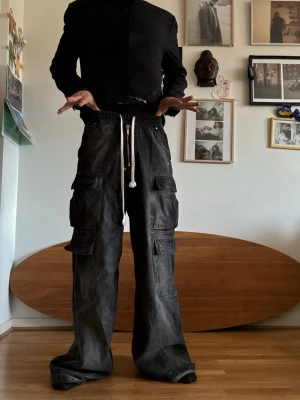 Vida svarta cargobyxor med snörning - Säljer ett par vida cargobyxor i svart denim med flera stora fickor på sidorna och vit dragsko i midjan. Byxorna har en avslappnad passform och streetwear-vibe, perfekta för dig som gillar oversized stil. Materialet är kraftigt och slitstarkt, troligen bomullsdenim.