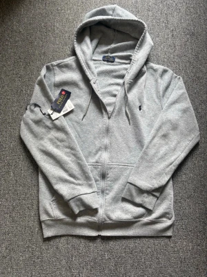 Grå hoodie från Polo Ralph Lauren - Snygg grå hoodie från Polo Ralph Lauren med dragkedja och huva. Klassisk logga broderad på bröstet och ribbade muddar. Mjuk bomullsblandning och praktiska fickor framtill. Perfekt för en avslappnad och cool look.