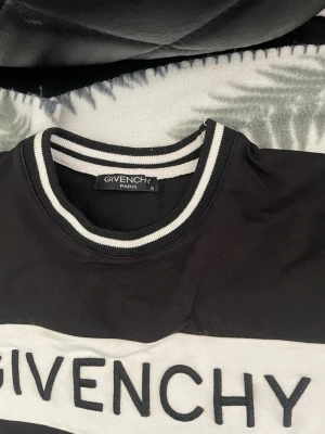 Svartvit sweatshirt från Givenchy - Säljer en svartvit sweatshirt från Givenchy med tydlig logga broderad över bröstet och röd Givenchy-symbol på magen. Tröjan har rund halsringning med svartvita ränder och tryck på ena ärmen. Perfekt för dig som gillar streetwear och exklusiva märken.